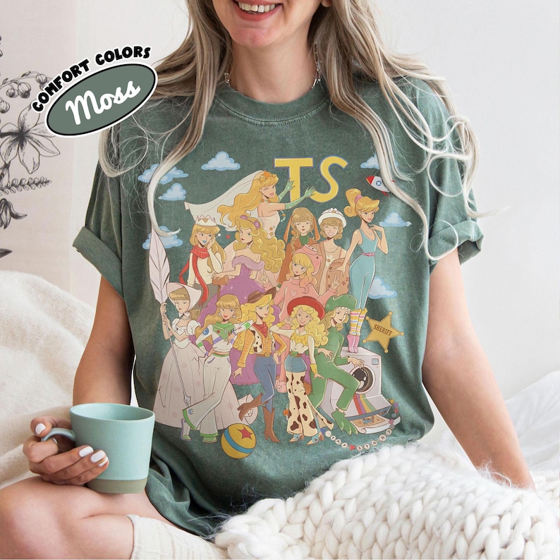 May include: A moss green t-shirt featuring a graphic print of multiple animated characters dressed in various costumes, including a space ranger, a cowgirl, and a sheriff. The text "TS" appears in yellow at the top, and a "Comfort Colors Moss" label is visible.