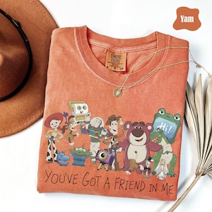 May include: A burnt orange crew neck t-shirt featuring a graphic print of various animated characters and the text "YOU'VE GOT A FRIEND IN ME" in black lettering. The shirt has a Comfort Colors label at the inner neckline and a short-sleeved design.