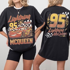 Może przedstawiać: Czarny t-shirt z motywem Zygzaka McQueena z filmu Auta. Prz&oacute;d koszulki zawiera numer 95, szachownicę i napis "McQueen". Tył koszulki r&oacute;wnież zawiera numer 95 i napis "Lightning McQueen".