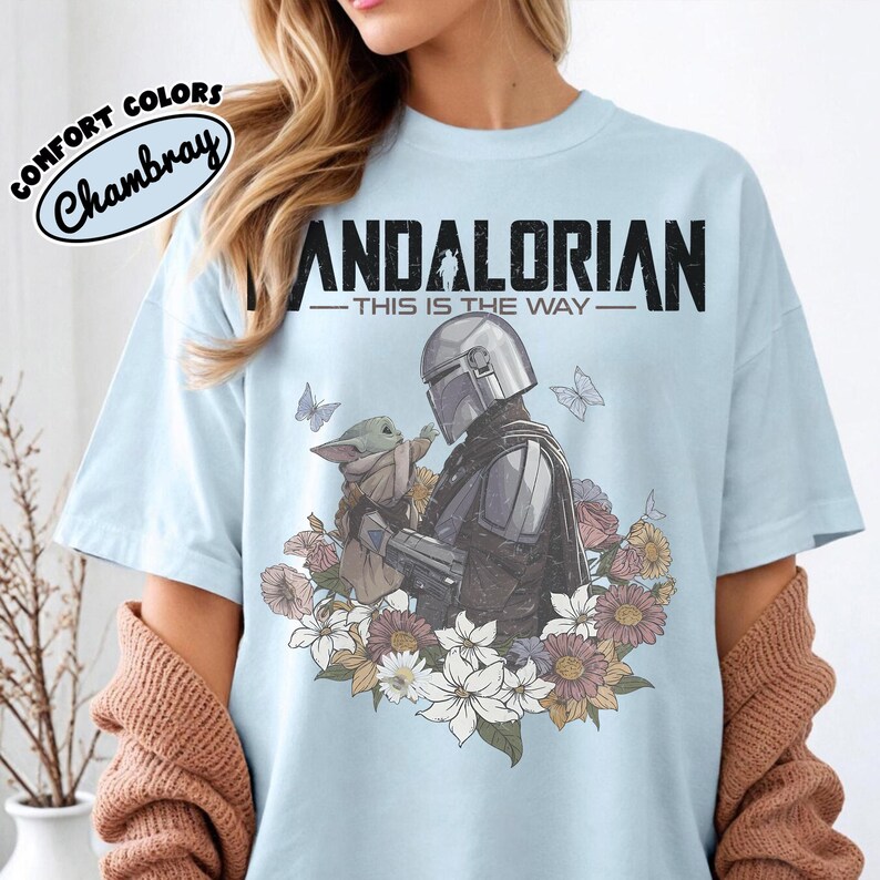 May include: Light blue t-shirt featuring a graphic of The Mandalorian and Grogu surrounded by flowers and butterflies. The text "MANDALORIAN - THIS IS THE WAY" is printed across the top. The shirt has a round neckline and short sleeves.