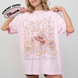 May include: A light pink t-shirt featuring a graphic print with the text Cowgirl Like Me and Takes One to Know One. The design includes a cowboy hat, roses, musical notes, and rope motifs in shades of brown, cream, and dark pink ink.