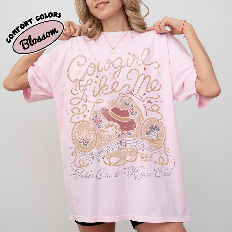 May include: A light pink t-shirt featuring a graphic print with the text Cowgirl Like Me and Takes One to Know One. The design includes a cowboy hat, roses, musical notes, and rope motifs in shades of brown, cream, and dark pink ink.