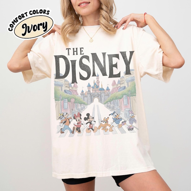Może przedstawiać: Kremowa koszulka z napisem "THE DISNEY" nad grafiką postaci Disneya przechodzących przez ulicę przed zamkiem. Koszulka ma wygląd vintage.