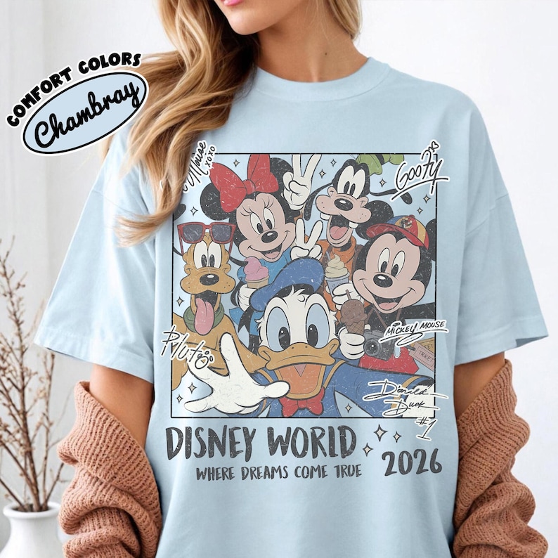 May include: Light blue t-shirt with a graphic of Disney characters, including Mickey Mouse, Minnie Mouse, and Goofy. The shirt has the text "DISNEY WORLD" and "2026" printed on it. The shirt is made of Chambray Comfort Colors material.