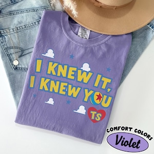 May include: A violet short-sleeved t-shirt featuring the yellow text I KNEW IT, I KNEW YOU with a red heart containing the letters TS, a colorful ball graphic, and white cloud and blue star accents printed on the front of the garment.