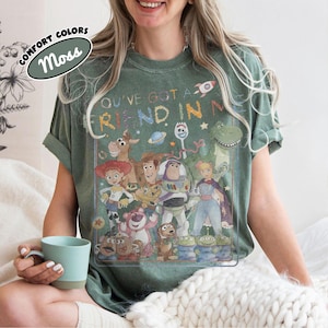 May include: A moss green t-shirt featuring a graphic print of various animated characters from the Toy Story franchise. The text "YOU'VE GOT A FRIEND IN ME" is printed above the characters in a colorful, distressed font. The shirt has a crew neck.