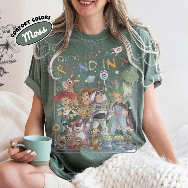 May include: A moss green t-shirt featuring a graphic print of various animated characters from the Toy Story franchise. The text "YOU'VE GOT A FRIEND IN ME" is printed above the characters in a colorful, distressed font. The shirt has a crew neck.