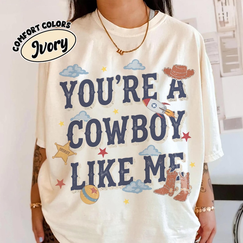 May include: An ivory t-shirt featuring the dark blue text "YOU'RE A COWBOY LIKE ME" surrounded by graphic illustrations of a brown cowboy hat, a rocket ship, a yellow sheriff star, a ball, and brown cowboy boots with blue clouds and stars.
