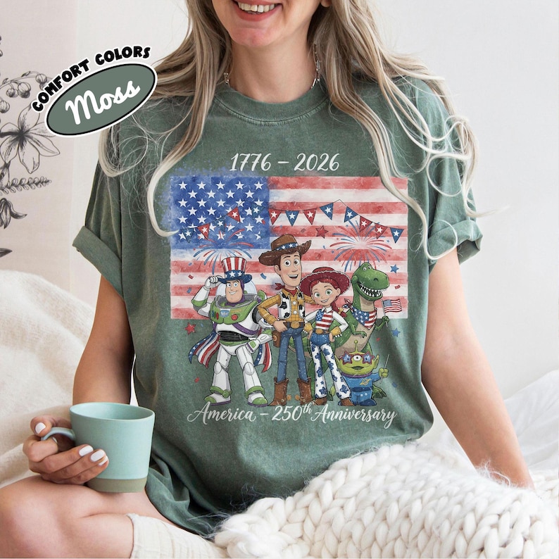 Może przedstawiać: Zielona koszulka z grafiką amerykańskiej flagi i postaciami z Toy Story. Koszulka zawiera napisy "1776-2026" i "America - 250th Anniversary". Koszulka jest w kolorze mchu.