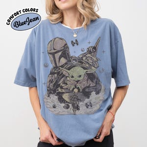 Puede incluir: Camiseta azul con un gr&aacute;fico que presenta personajes de una popular serie de ciencia ficci&oacute;n. El dise&ntilde;o incluye una figura con casco, una peque&ntilde;a criatura verde y una nave espacial. La camiseta tiene el texto "Comfort Colors Blue Jean" en un dise&ntilde;o circular.