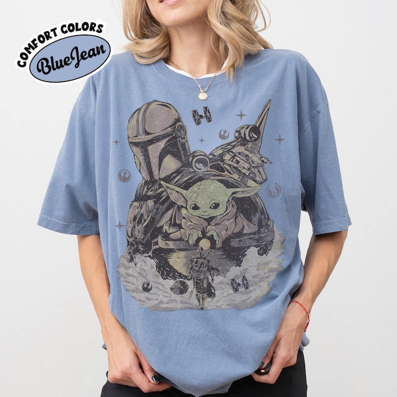 Puede incluir: Camiseta azul con un gr&aacute;fico que presenta personajes de una popular serie de ciencia ficci&oacute;n. El dise&ntilde;o incluye una figura con casco, una peque&ntilde;a criatura verde y una nave espacial. La camiseta tiene el texto "Comfort Colors Blue Jean" en un dise&ntilde;o circular.