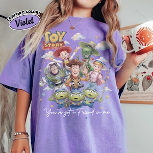 May include: A violet Comfort Colors t-shirt with a Toy Story character graphic. The design features Woody, Buzz Lightyear, Jessie, Rex, Bo Peep, and the Little Green Men. The text "You've got a Friend in me" is also included.