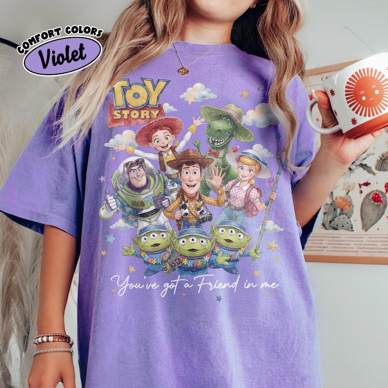 May include: A violet Comfort Colors t-shirt with a Toy Story character graphic. The design features Woody, Buzz Lightyear, Jessie, Rex, Bo Peep, and the Little Green Men. The text "You've got a Friend in me" is also included.