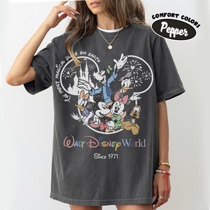 May include: Dark gray t-shirt featuring a colorful graphic of Disney characters, including Mickey Mouse, Minnie Mouse, and Donald Duck. The shirt has the text "Walt Disney World Since 1971" and "The Most Magical Place on Earth".