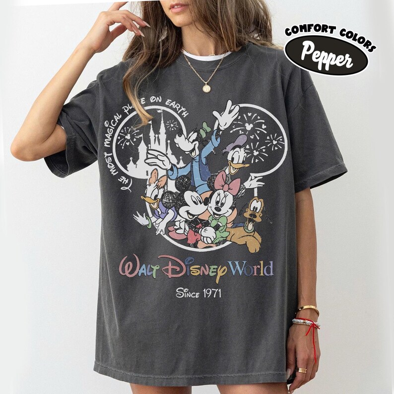 May include: Dark gray t-shirt featuring a colorful graphic of Disney characters, including Mickey Mouse, Minnie Mouse, and Donald Duck. The shirt has the text "Walt Disney World Since 1971" and "The Most Magical Place on Earth".