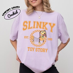 Pu&ograve; includere: T-shirt Orchid Comfort Colors con grafica di Slinky Dog di Toy Story. Il design include la parola "SLINKY" in lettere arancioni, il testo "EST. 1995" e le parole "TOY STORY".