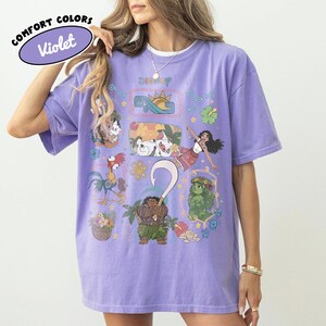 May include: A light purple Violet Comfort Colors t-shirt featuring Disney characters. The shirt has a crew neck and short sleeves. The graphic includes illustrations of characters and the word "Disney".