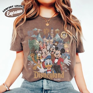 May include: Espresso-colored t-shirt featuring a Disneyland graphic with Mickey Mouse, Minnie Mouse, Donald Duck, Goofy, and park scenery. The text "Disneyland Est.1955" is printed below the characters. The shirt has short sleeves and a crew neck.