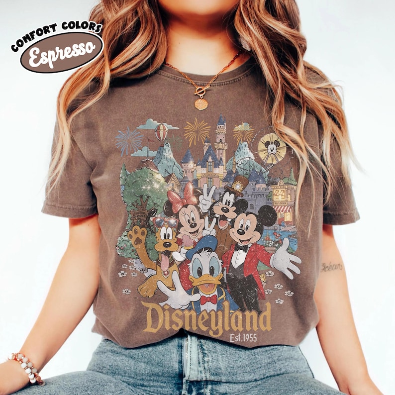 May include: Espresso-colored t-shirt featuring a Disneyland graphic with Mickey Mouse, Minnie Mouse, Donald Duck, Goofy, and park scenery. The text "Disneyland Est.1955" is printed below the characters. The shirt has short sleeves and a crew neck.
