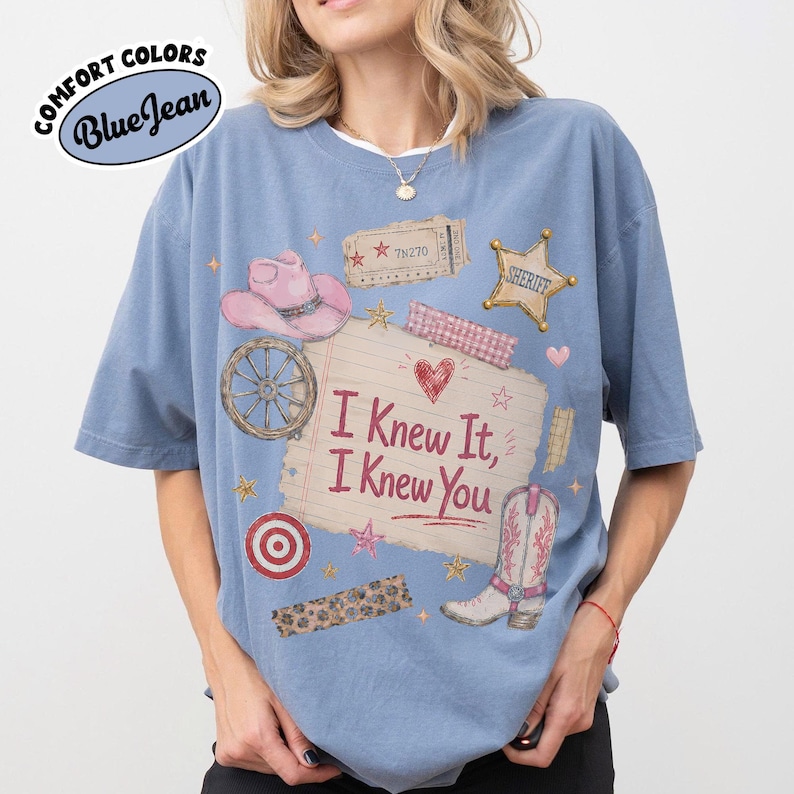 Puede incluir: Una camiseta azul de manga corta con un estampado gr&aacute;fico de un sombrero de vaquero rosa, una rueda de carreta, una estrella de sheriff, una bota de vaquero y un papel rayado con el texto I Knew It, I Knew You, decorado con estrellas y un coraz&oacute;n.