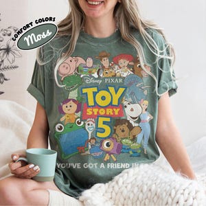 Op de afbeelding: Een mosgroen T-shirt met korte mouwen en een grafische print van diverse Toy Story-personages, het Disney Pixar-logo, de tekst Toy Story 5 en de zin You've Got A Friend In Me. Het T-shirt heeft een vervaagde uitstraling in vintage-stijl.
