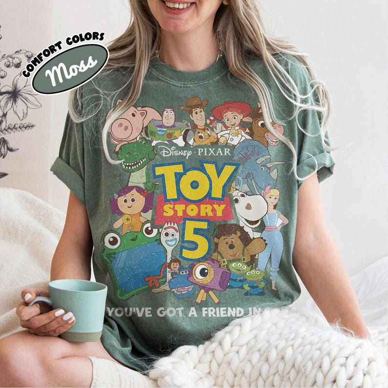 Op de afbeelding: Een mosgroen T-shirt met korte mouwen en een grafische print van diverse Toy Story-personages, het Disney Pixar-logo, de tekst Toy Story 5 en de zin You've Got A Friend In Me. Het T-shirt heeft een vervaagde uitstraling in vintage-stijl.