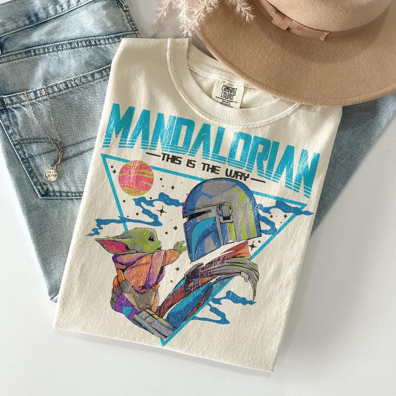 Może przedstawiać: Kremowy t-shirt z grafiką Mandalorian i Grogu, z napisem "MANDALORIAN" w kolorze turkusowym. Projekt zawiera tr&oacute;jkąt z r&oacute;żową i ż&oacute;łtą planetą. Koszulka jest złożona na niebieskich dżinsach, z naszyjnikiem i beżowym kapeluszem.