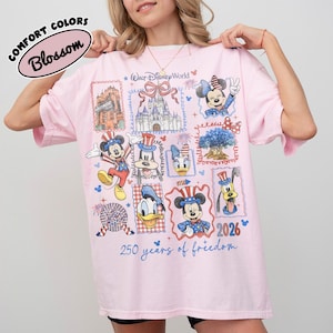 May include: A light pink t-shirt featuring a colorful graphic design with various Disney characters and landmarks. The design includes Mickey Mouse, Minnie Mouse, Donald Duck, and Pluto, along with the text "Walt Disney World" and "250 years of freedom".