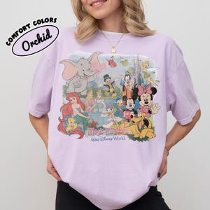 May include: A light purple t-shirt featuring a collage of Disney characters, including Dumbo, Ariel, Mickey Mouse, and Minnie Mouse. The text "Magic Kingdom Walt Disney World" is printed below the characters. The shirt has the text "COMFORT COLORS Orchid" in a bubble.