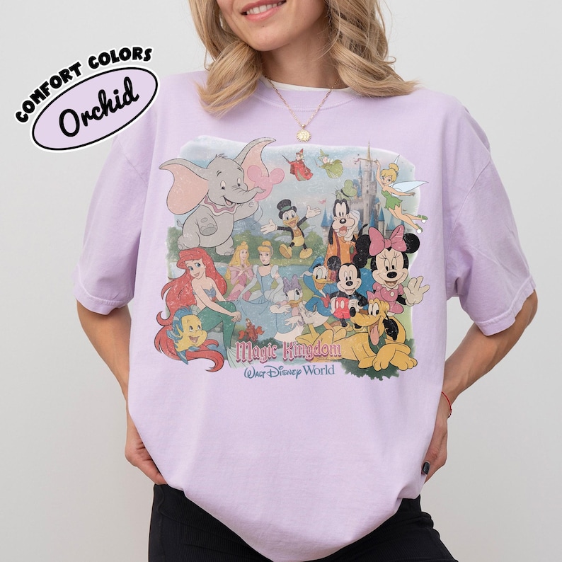 May include: A light purple t-shirt featuring a collage of Disney characters, including Dumbo, Ariel, Mickey Mouse, and Minnie Mouse. The text "Magic Kingdom Walt Disney World" is printed below the characters. The shirt has the text "COMFORT COLORS Orchid" in a bubble.
