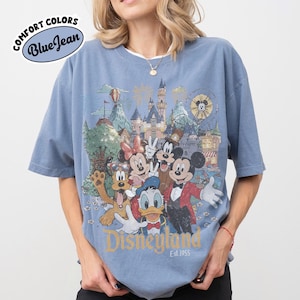 May include: Blue t-shirt with a Disneyland graphic featuring Mickey Mouse, Minnie Mouse, Donald Duck, Goofy, and other characters. The shirt has the text "Disneyland Est. 1955" and "Comfort Colors Blue Jean" on the top left.