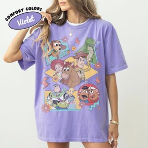 Puede incluir: Una camiseta violeta con un gr&aacute;fico de personajes de la pel&iacute;cula animada Toy Story. La camiseta incluye el texto "COMFORT COLORS" y "Violet" en un dise&ntilde;o circular. El gr&aacute;fico incluye im&aacute;genes de Woody, Buzz Lightyear y otros personajes.