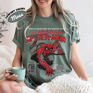 May include: A moss green short-sleeve t-shirt featuring a vintage-style graphic of Spider-Man in red and black. The text reads: Never has anyone fought such merciless foes, The Amazing Spider-Man, Peter Parker, and Another spectacular smash-hit.