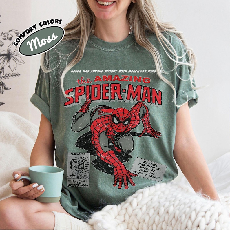 May include: A moss green short-sleeve t-shirt featuring a vintage-style graphic of Spider-Man in red and black. The text reads: Never has anyone fought such merciless foes, The Amazing Spider-Man, Peter Parker, and Another spectacular smash-hit.