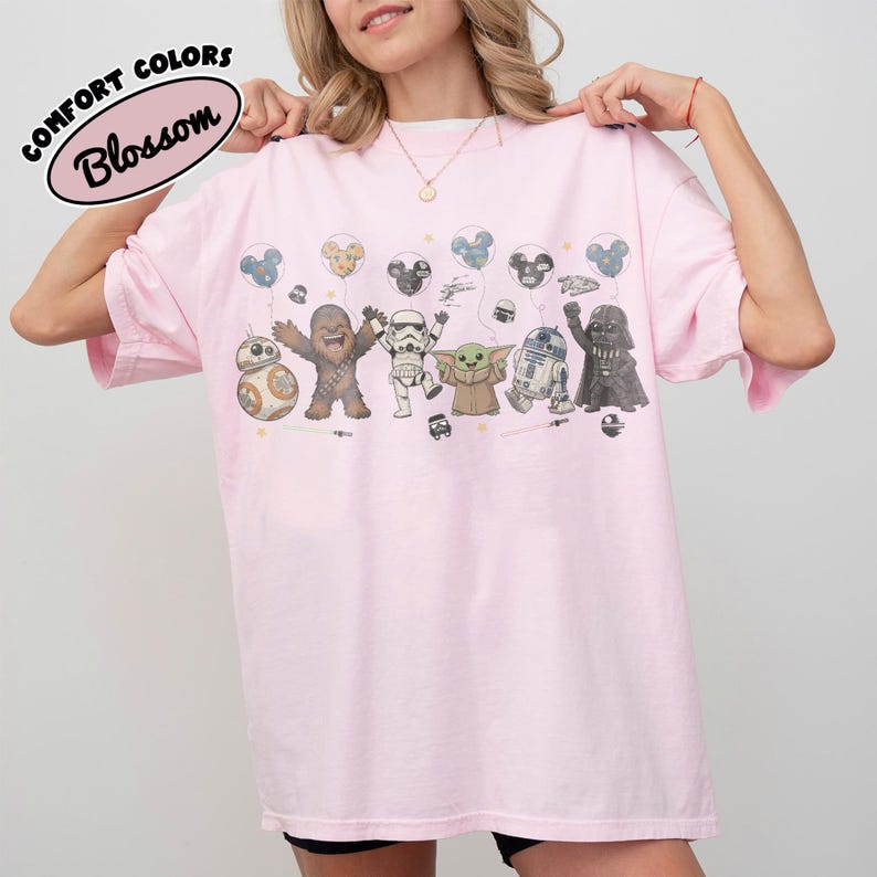 K&ouml;nnte beinhalten: Ein hellrosa T-Shirt mit einem Grafikdruck von Star Wars-Figuren, darunter BB-8, Chewbacca, ein Stormtrooper, Grogu, R2-D2 und Darth Vader, die Mickey-Maus-f&ouml;rmige Luftballons halten. Der Text Comfort Colors Blossom steht oben links im Bild.