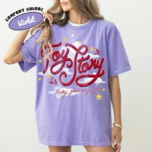 May include: A violet short-sleeve t-shirt featuring a graphic design with the red rope-style text Toy Story, white clouds, yellow stars, and small red flowers. Below the main text, the phrase Baby, just say yes is printed in red cursive script.