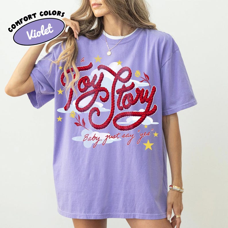 May include: A violet short-sleeve t-shirt featuring a graphic design with the red rope-style text Toy Story, white clouds, yellow stars, and small red flowers. Below the main text, the phrase Baby, just say yes is printed in red cursive script.