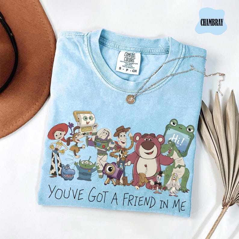 May include: A light blue crew neck t-shirt featuring a graphic print of various Toy Story characters including Woody, Buzz Lightyear, Jessie, and Lotso. The text "YOU'VE GOT A FRIEND IN ME" is printed in black below the characters. The shirt has a Comfort Colors tag.