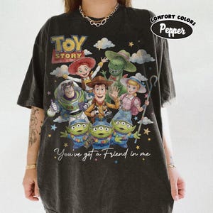 May include: Black Comfort Colors t-shirt with a Toy Story graphic. The design includes Woody, Buzz Lightyear, and the Little Green Men. The phrase "You've got a Friend in me" is printed below the characters.