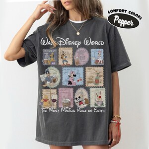 May include: Dark gray t-shirt featuring a Walt Disney World design with various Disney characters and country stamps. The text "The Most Magical Place on Earth" is printed below the stamps. The shirt also has the text "Comfort Colors Pepper".