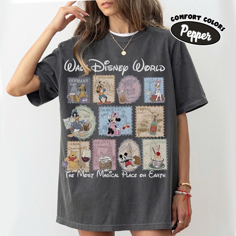 May include: Dark gray t-shirt featuring a Walt Disney World design with various Disney characters and country stamps. The text "The Most Magical Place on Earth" is printed below the stamps. The shirt also has the text "Comfort Colors Pepper".