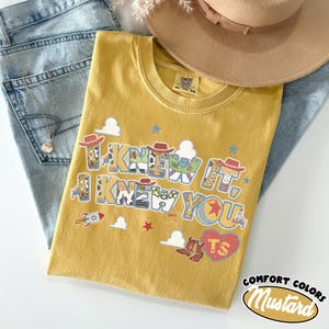 May include: A mustard yellow t-shirt featuring the text "I KNEW IT, I KNEW YOU" in a bold font. The letters are filled with patterns of cowboy hats, boots, and space ranger suits. Small graphics of rockets, stars, and clouds surround the text.