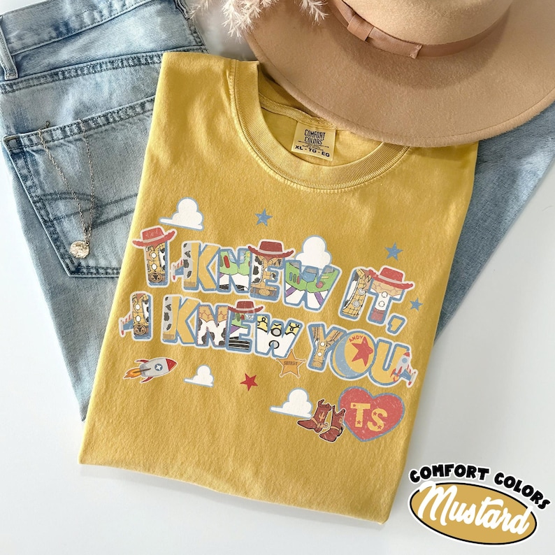 May include: A mustard yellow t-shirt featuring the text "I KNEW IT, I KNEW YOU" in a bold font. The letters are filled with patterns of cowboy hats, boots, and space ranger suits. Small graphics of rockets, stars, and clouds surround the text.
