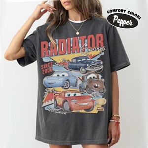 May include: Dark gray t-shirt featuring a graphic of characters from the movie Cars, including Lightning McQueen, Mater, and Doc Hudson. The shirt has the words "Radiator Springs" and "Since 2006" printed on it. The shirt is a Comfort Colors Pepper tee.