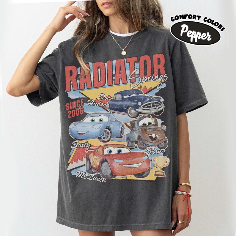 May include: Dark gray t-shirt featuring a graphic of characters from the movie Cars, including Lightning McQueen, Mater, and Doc Hudson. The shirt has the words "Radiator Springs" and "Since 2006" printed on it. The shirt is a Comfort Colors Pepper tee.