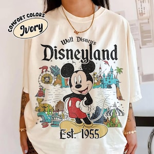 Op de afbeelding: Een ivoorkleurig t-shirt met een grafische print van Mickey Mouse gecentreerd boven de tekst Est. 1955. Het ontwerp bevat illustraties van Disneyland-attracties, een kasteel en de tekst Walt Disney's Disneyland in een verweerd zwart lettertype.