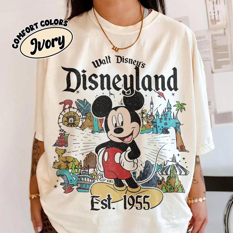 Op de afbeelding: Een ivoorkleurig t-shirt met een grafische print van Mickey Mouse gecentreerd boven de tekst Est. 1955. Het ontwerp bevat illustraties van Disneyland-attracties, een kasteel en de tekst Walt Disney's Disneyland in een verweerd zwart lettertype.