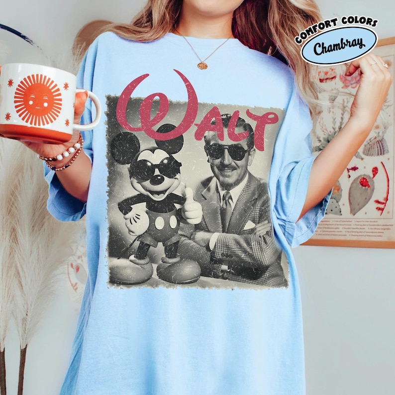 K&ouml;nnte beinhalten: Hellblaues T-Shirt mit einem Vintage-Grafik von Mickey Mouse und Walt Disney, mit dem Wort "WALT" in Rot. Das Shirt hat eine lockere Passform und die Worte "COMFORT COLORS" und "Chambray" sind oben rechts aufgedruckt.