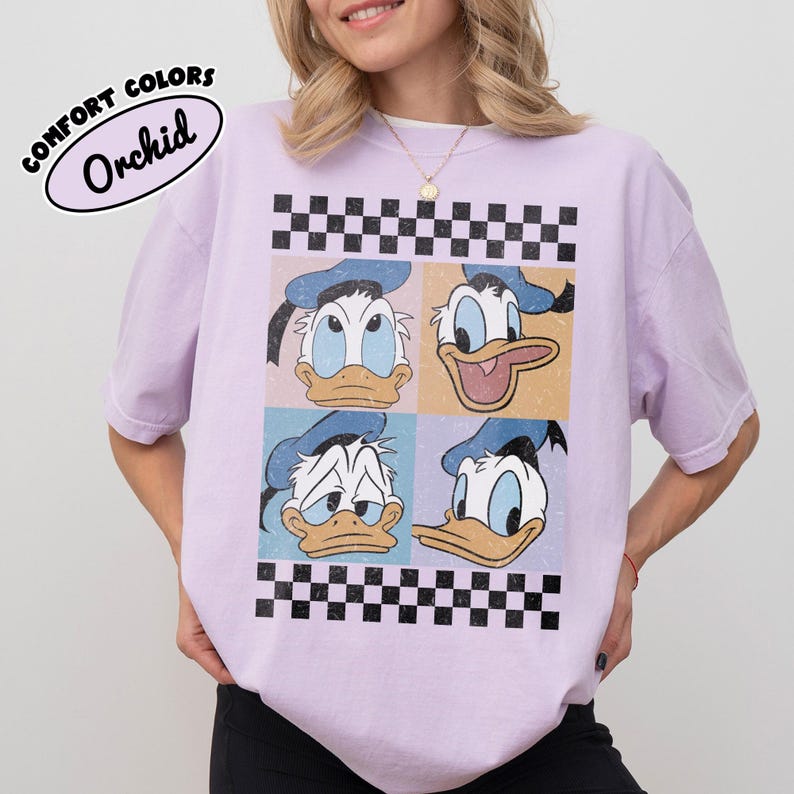 May include: A light purple t-shirt featuring a graphic of Donald Duck's face in four different expressions, set against a checkered background. The text "COMFORT COLORS Orchid" is printed on the upper left.
