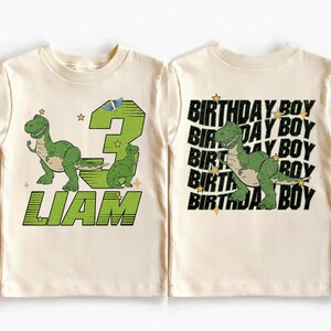 K&ouml;nnte beinhalten: Cremefarbenes Kinder-T-Shirt mit einem gr&uuml;nen Dinosaurier-Motiv. Auf der Vorderseite sind die Zahl 3, der Name "LIAM" und eine Dinosaurier-Illustration zu sehen. Auf der R&uuml;ckseite wiederholt sich "BIRTHDAY BOY" mit einer Dinosaurier-Grafik.